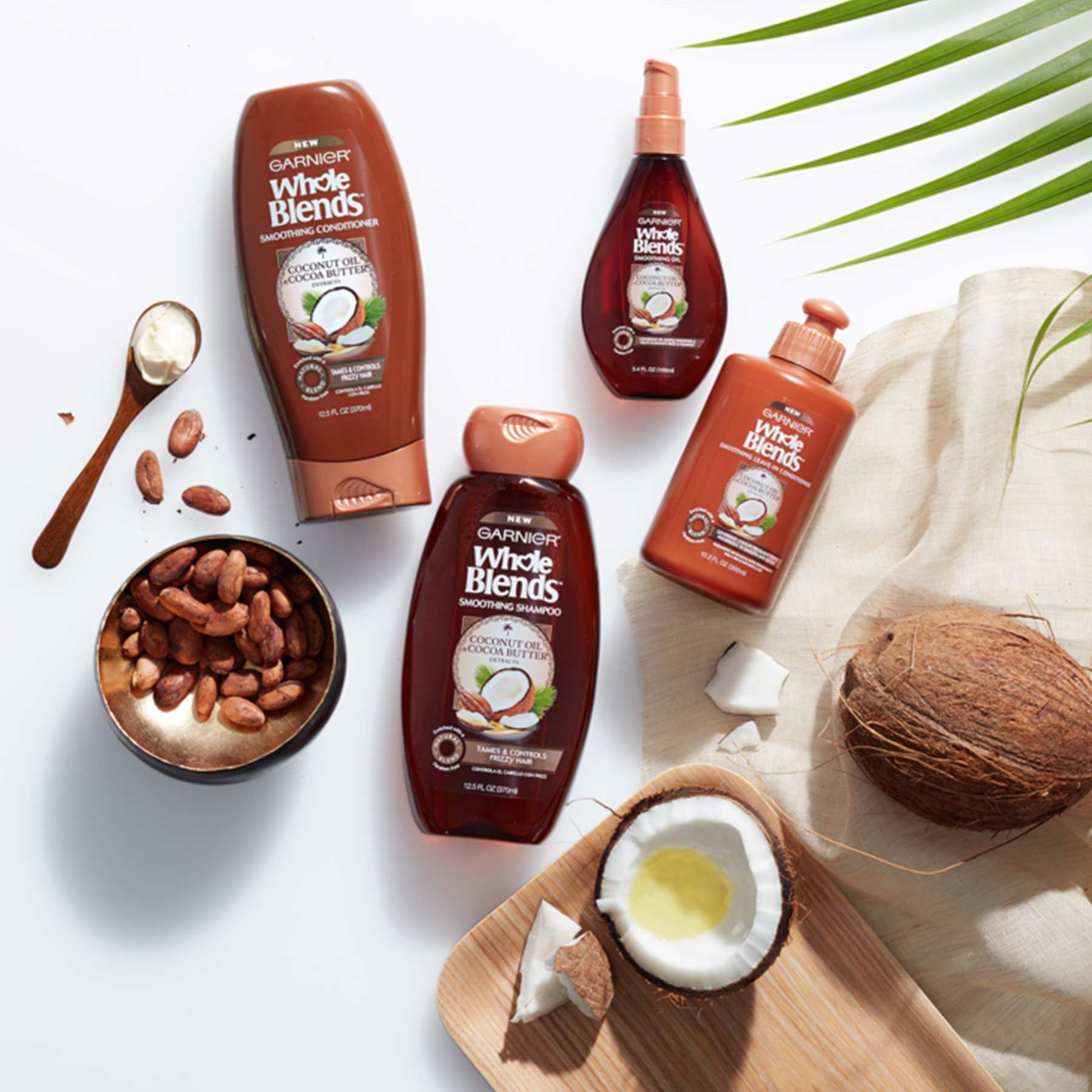 Whole Blends Coconut Oil and Cocoa Butter Shampoo, Coconut Oil and Cocoa Butter Conditioner, Coconut Oil and Cocoa Butter Leave-in Conditioner, and Blends Coconut Oil and Cocoa Butter Smoothing Oil next to a wooden bowl of cocoa beans with some spilled out, a wooden spoon of cocoa butter, a wooden tray with a broken coconut with coconut oil puddled in the halved piece, a natural-colored linen with a coconut, and a palm frond on a white background.